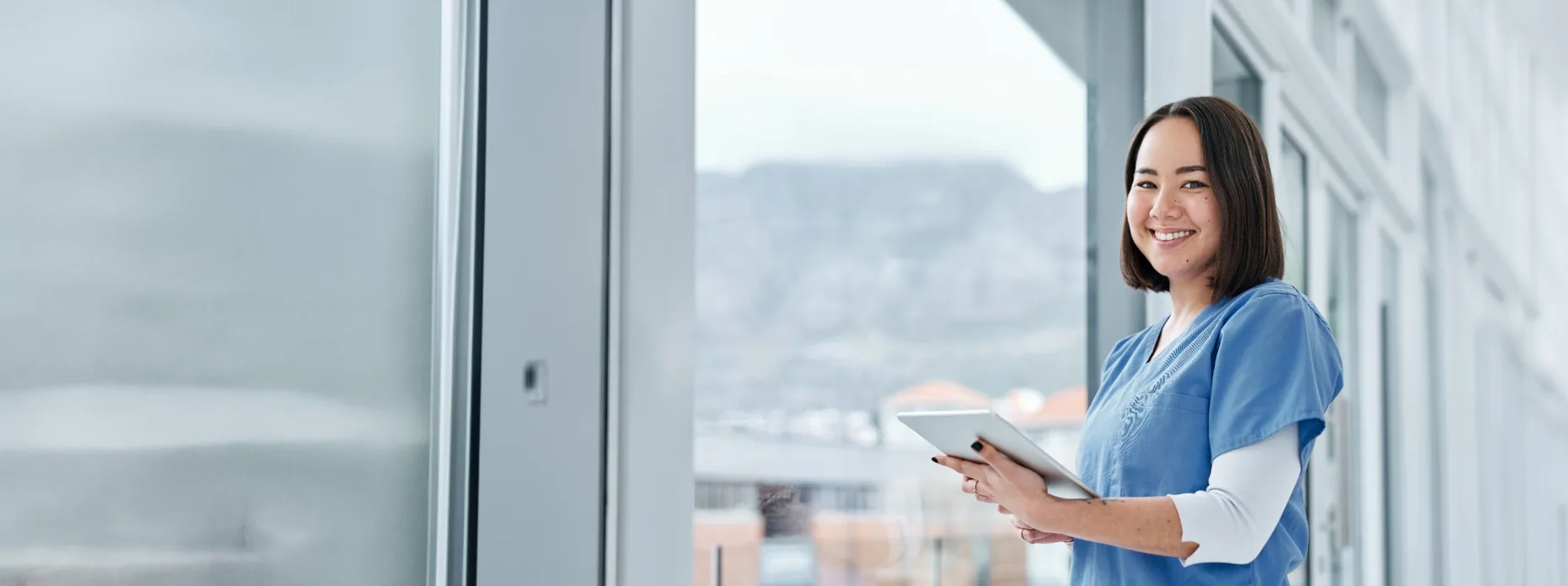 Nurse in blue scrubs holding a tablet in a bright hospital corridor