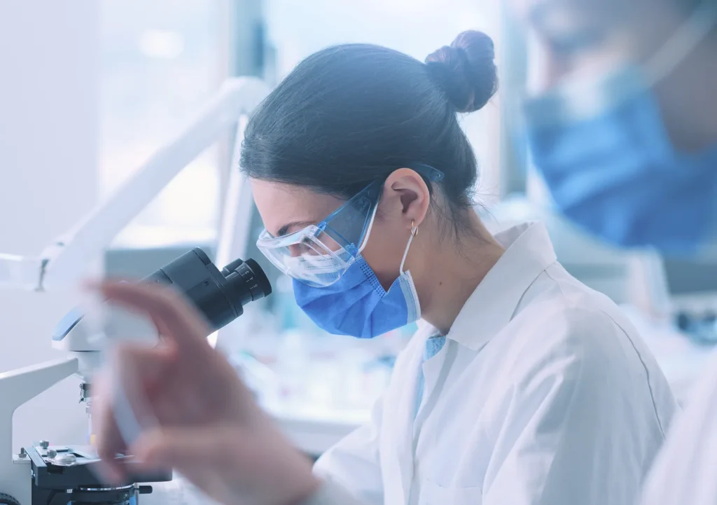Researcher in a mask and safety glasses working at a microscope in a laboratory