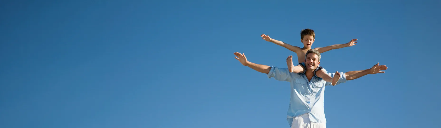Father carrying a child on his shoulders, both with arms outstretched against a blue sky